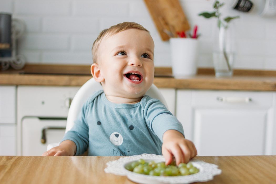 pinzettengriff meilenstein der feinmotorik, froehliches baby greift traube beim essen im hochstuhl