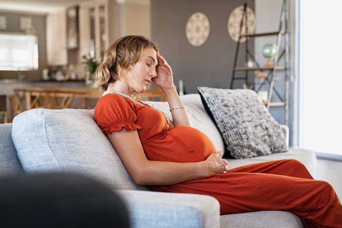 schwangere frau mit kopfschmerzen in der schwangerschaft sitzt auf sofa hand an kopf und babybauch